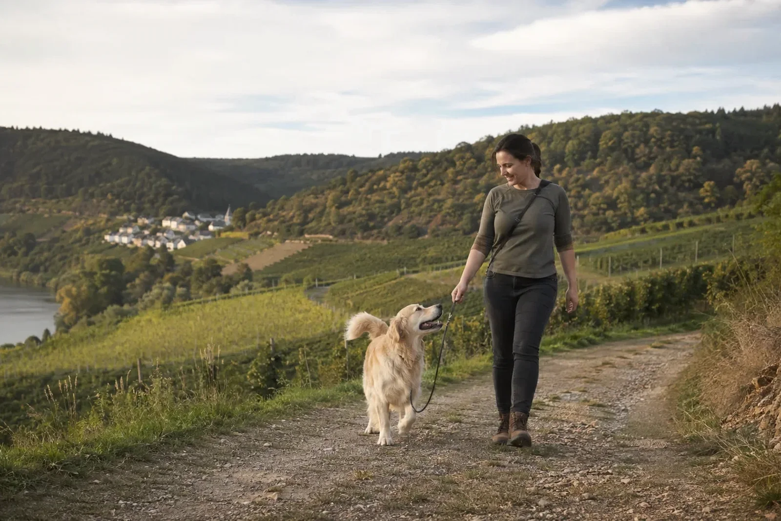 Eine Frau geht mit einem Golden Retriever an lockerer Leine auf einem Weg oberhalb eines Flusses.