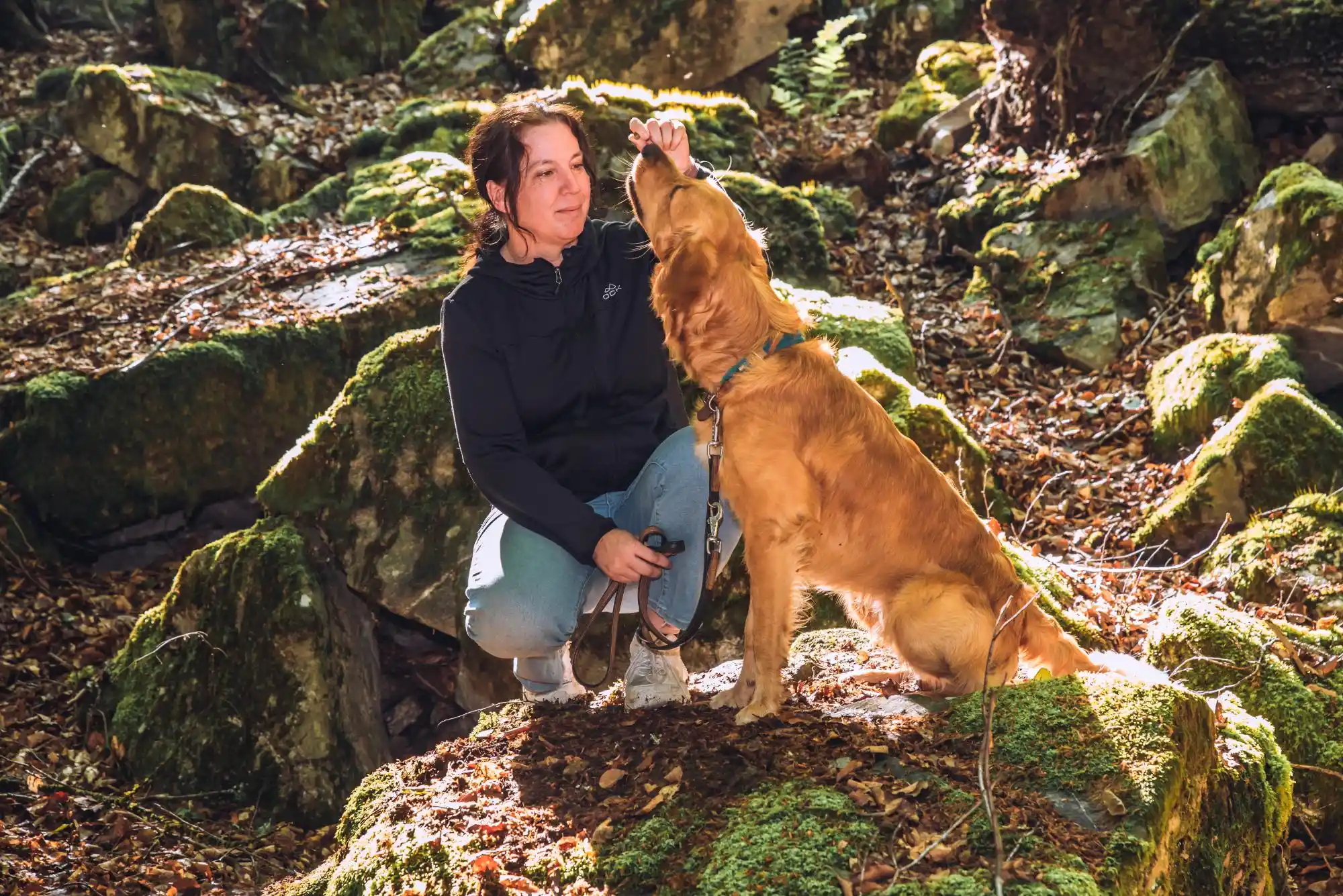 Ein Golden Retriever streckt seine Schnauze zu Kerstin Ehses, die neben ihm auf einem moosigen Stein hockt.