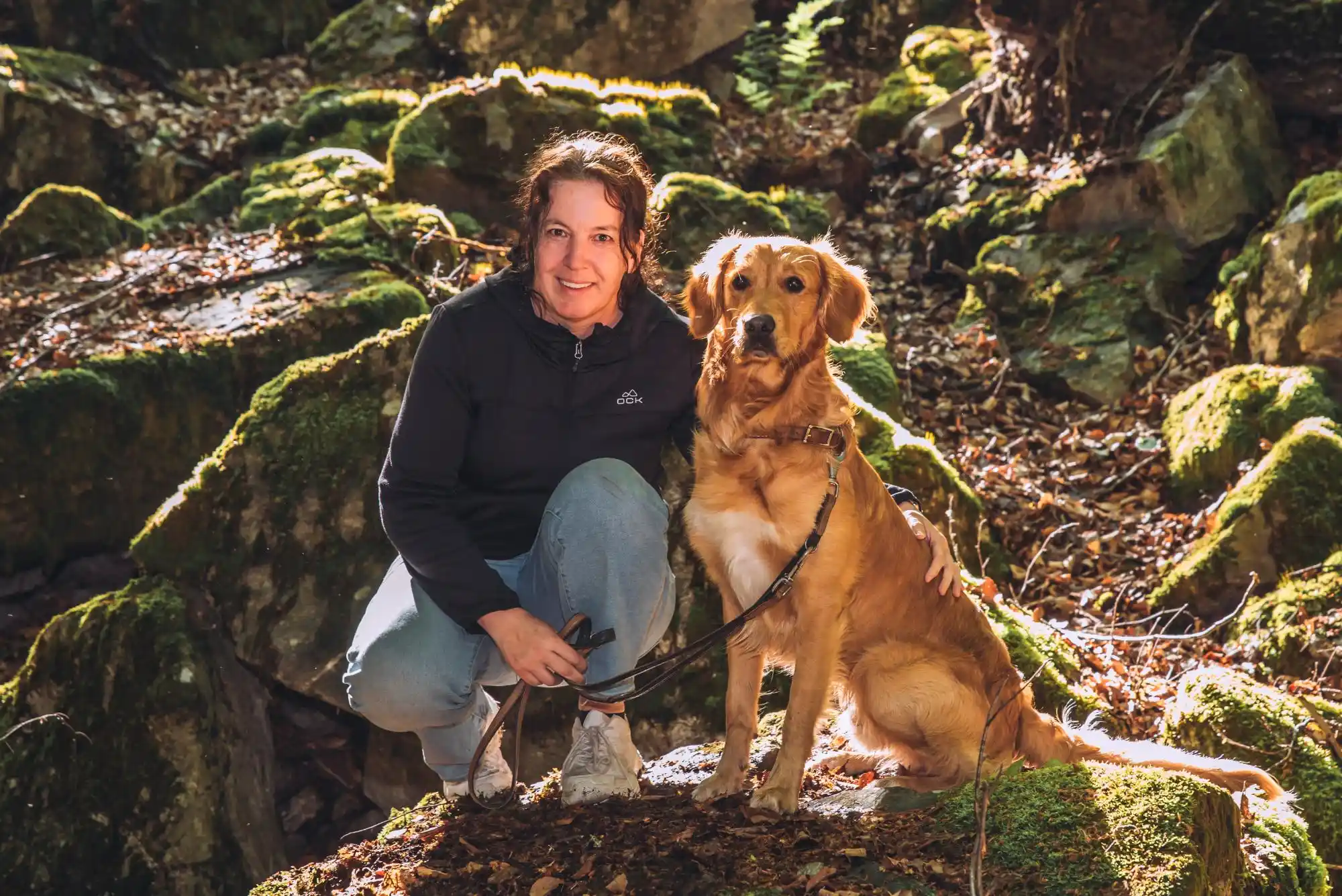 Kerstin Ehses hockt neben einem sitzenden Golden Retriever auf moosigen Steinen und blickt zur Kamera.